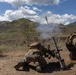 Two military personnel in full combat gear are operating a mortar system, with a round visibly in flight just after firing. The scene takes place in an open, arid training or operational area with green hills and mountains in the background. The image captures the moment of discharge with visible propellant smoke rising from the mortar tube.