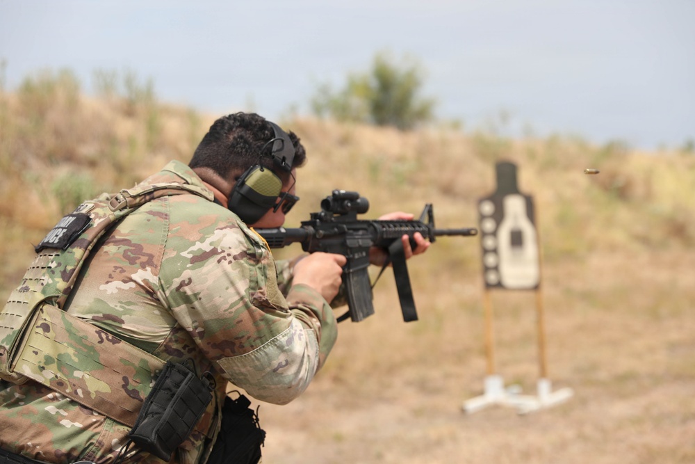 A military or law enforcement personnel is engaged in a live-fire shooting exercise at an outdoor range. The shooter is in a crouched stance aiming an M4-style carbine rifle at a silhouette target downrange. A spent brass casing is visibly ejecting from the weapon, indicating the moment of firing.