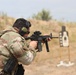 A military or law enforcement personnel is engaged in a live-fire shooting exercise at an outdoor range. The shooter is in a crouched stance aiming an M4-style carbine rifle at a silhouette target downrange. A spent brass casing is visibly ejecting from the weapon, indicating the moment of firing.