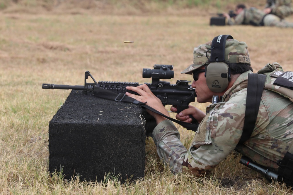 A soldier in MultiCam/OCP camouflage uniform is in a prone firing position on a grass range, aiming an M4/M16-style rifle rested on a black foam block. A spent brass casing is visibly ejecting from the weapon, indicating the rifle has just been fired. Additional personnel in similar uniforms are visible in the background, also in prone positions, suggesting a organized military marksmanship training exercise.