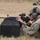 Law Enforcement Agency Conducts LEWTAQ at Fort Hood A soldier in MultiCam/OCP camouflage uniform is in a prone firing position on a grass range, aiming an M4/M16-style rifle rested on a black foam block. A spent brass casing is visibly ejecting from the weapon, indicating the rifle has just been fired. Additional personnel in similar uniforms are visible in the background, also in prone positions, suggesting a organized military marksmanship training exercise.
