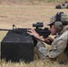 A soldier in MultiCam/OCP camouflage uniform is in a prone firing position on a grass range, aiming an M4/M16-style rifle rested on a black foam block. A spent brass casing is visibly ejecting from the weapon, indicating the rifle has just been fired. Additional personnel in similar uniforms are visible in the background, also in prone positions, suggesting a organized military marksmanship training exercise.