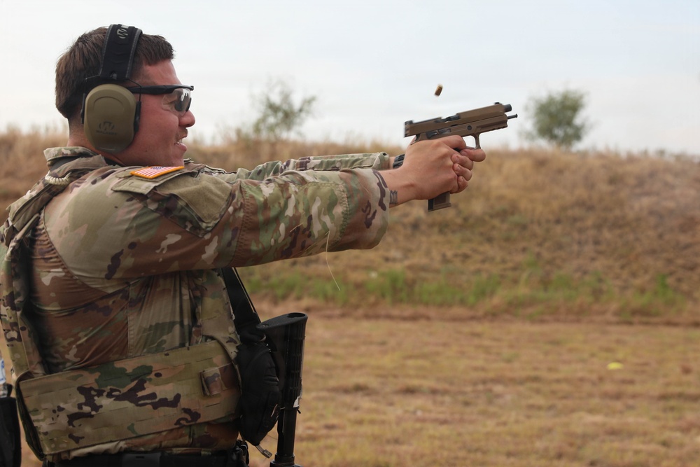 A U.S. Army soldier in MultiCam (OCP) camouflage uniform is firing a pistol at an outdoor shooting range. A spent brass casing is visibly ejecting from the firearm mid-shot. The soldier is wearing proper hearing and eye protection, indicating a controlled training or competition environment.