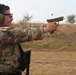 A U.S. Army soldier in MultiCam (OCP) camouflage uniform is firing a pistol at an outdoor shooting range. A spent brass casing is visibly ejecting from the firearm mid-shot. The soldier is wearing proper hearing and eye protection, indicating a controlled training or competition environment.