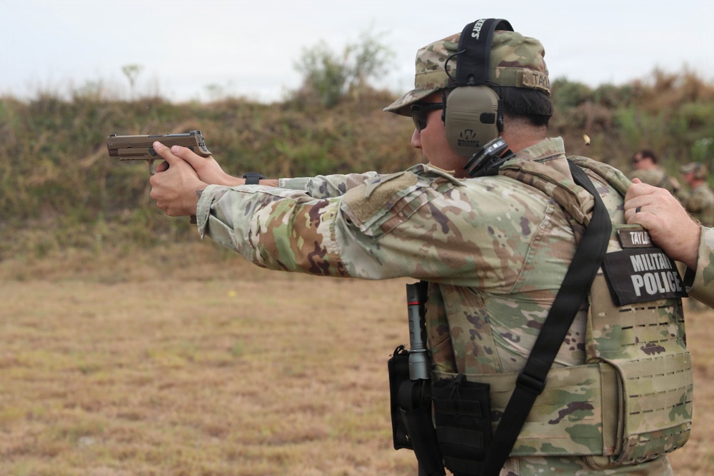 A U.S. Army Military Police soldier named Taylor is firing a pistol at a shooting range. The soldier is wearing full MultiCam (OCP) uniform with a plate carrier vest, hearing protection, and a patrol cap. A second individual is visible behind the shooter, appearing to provide oversight or instruction.