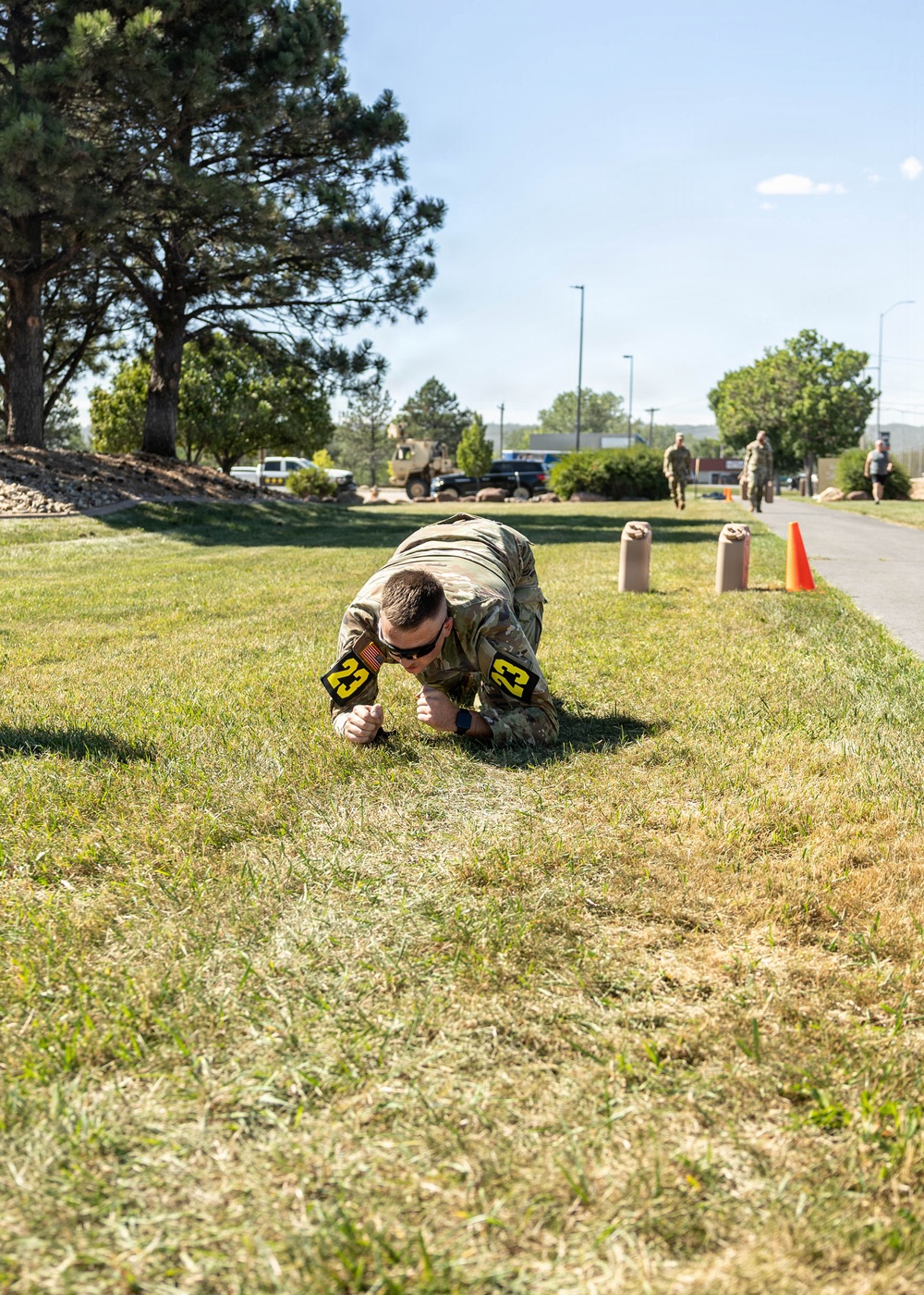 A soldier in camouflage uniform is performing a low crawl exercise on a grass field, wearing number '23' identification bands on both arms. The activity appears to be part of a military fitness competition or training event, with sandbags and an orange traffic cone visible as course markers in the background. Additional military personnel and vehicles are visible in the background along a paved path.