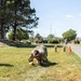 A soldier in camouflage uniform is performing a low crawl exercise on a grass field, wearing number '23' identification bands on both arms. The activity appears to be part of a military fitness competition or training event, with sandbags and an orange traffic cone visible as course markers in the background. Additional military personnel and vehicles are visible in the background along a paved path.