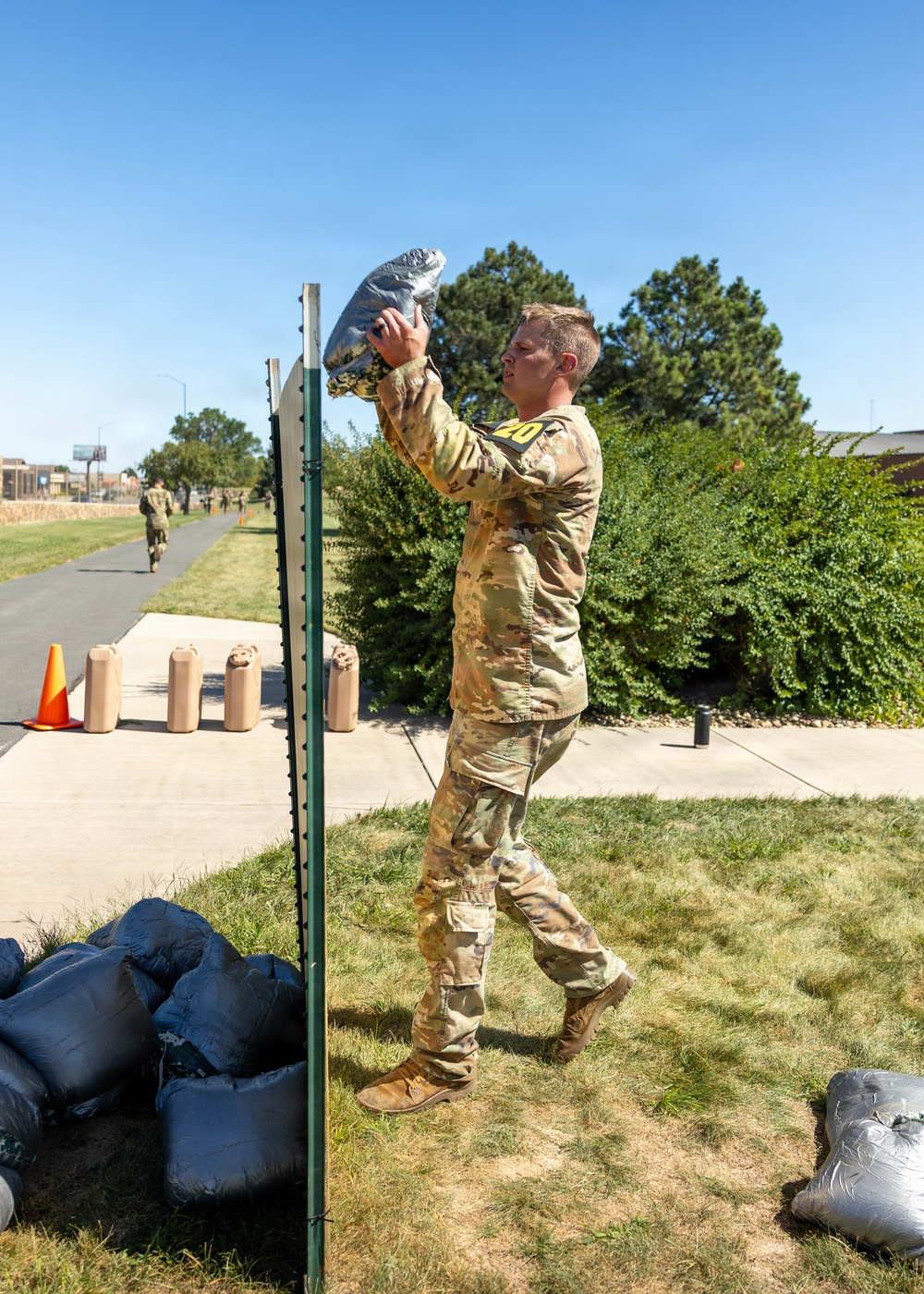 A soldier in OCP (Operational Camouflage Pattern) uniform, wearing a numbered bib '20', is lifting a weighted sandbag or filled bag overhead near a vertical post or height marker. The activity appears to be part of a military fitness competition or physical assessment event, taking place outdoors on a military installation on a clear sunny day. Additional soldiers and orange traffic cones are visible in the background along a paved path.
