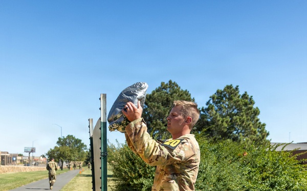 A soldier in OCP (Operational Camouflage Pattern) uniform, wearing a numbered bib '20', is lifting a weighted sandbag or filled bag overhead near a vertical post or height marker. The activity appears to be part of a military fitness competition or physical assessment event, taking place outdoors on a military installation on a clear sunny day. Additional soldiers and orange traffic cones are visible in the background along a paved path.