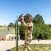 A soldier in OCP (Operational Camouflage Pattern) uniform, wearing a numbered bib '20', is lifting a weighted sandbag or filled bag overhead near a vertical post or height marker. The activity appears to be part of a military fitness competition or physical assessment event, taking place outdoors on a military installation on a clear sunny day. Additional soldiers and orange traffic cones are visible in the background along a paved path.