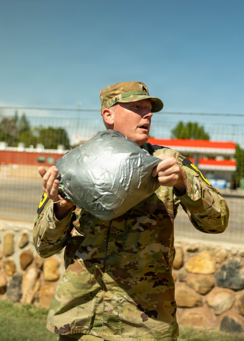 A U.S. Army soldier in OCP (Operational Camouflage Pattern) uniform is holding a large silver duct-tape-wrapped bundle or package. The soldier appears to be outdoors in a sunny environment, possibly on or near a military installation or urban area. Yellow numbered markers are visible on the soldier's sleeves, suggesting a training exercise or competition event.