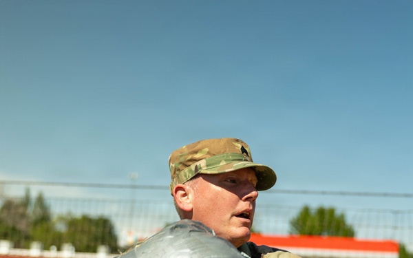 A U.S. Army soldier in OCP (Operational Camouflage Pattern) uniform is holding a large silver duct-tape-wrapped bundle or package. The soldier appears to be outdoors in a sunny environment, possibly on or near a military installation or urban area. Yellow numbered markers are visible on the soldier's sleeves, suggesting a training exercise or competition event.