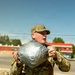 A U.S. Army soldier in OCP (Operational Camouflage Pattern) uniform is holding a large silver duct-tape-wrapped bundle or package. The soldier appears to be outdoors in a sunny environment, possibly on or near a military installation or urban area. Yellow numbered markers are visible on the soldier's sleeves, suggesting a training exercise or competition event.