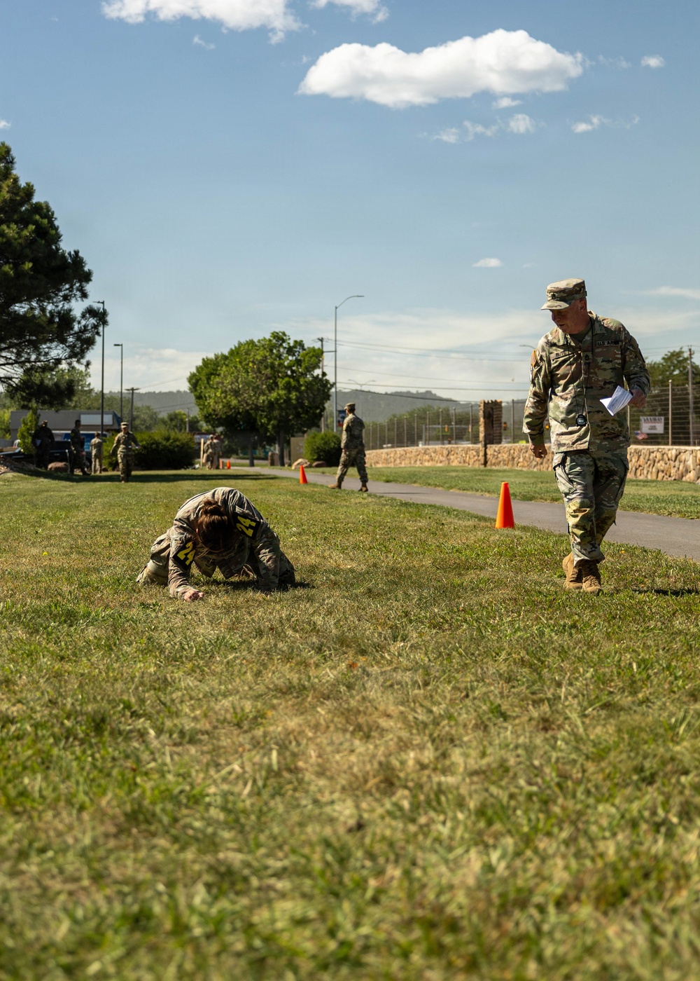 A military fitness or assessment event taking place on a grassy field at what appears to be a U.S. Army installation. One soldier wearing a numbered bib (number 24) is in a low crawl or collapsed position on the grass, while a supervising soldier in OCP uniform holds papers and observes. Orange traffic cones mark a course route in the background.