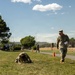 A military fitness or assessment event taking place on a grassy field at what appears to be a U.S. Army installation. One soldier wearing a numbered bib (number 24) is in a low crawl or collapsed position on the grass, while a supervising soldier in OCP uniform holds papers and observes. Orange traffic cones mark a course route in the background.