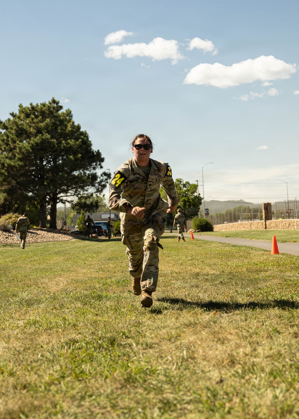 A military service member wearing camouflage uniform and number '24' bib markers on both arms is running across a grassy area, appearing to be participating in a timed fitness or competition event. The setting appears to be a military installation with other uniformed personnel visible in the background. An orange traffic cone marks the course route to the right.
