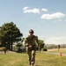 A military service member wearing camouflage uniform and number '24' bib markers on both arms is running across a grassy area, appearing to be participating in a timed fitness or competition event. The setting appears to be a military installation with other uniformed personnel visible in the background. An orange traffic cone marks the course route to the right.