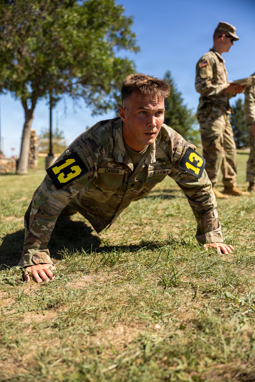 A U.S. Army soldier identified by name tape as 'CARDA' and wearing number '13' bib patches on both shoulders performs push-ups on a grass field. A second uniformed soldier stands in the background holding a clipboard or tablet, apparently observing and scoring the performance. The setting appears to be an outdoor physical fitness test or military competition.