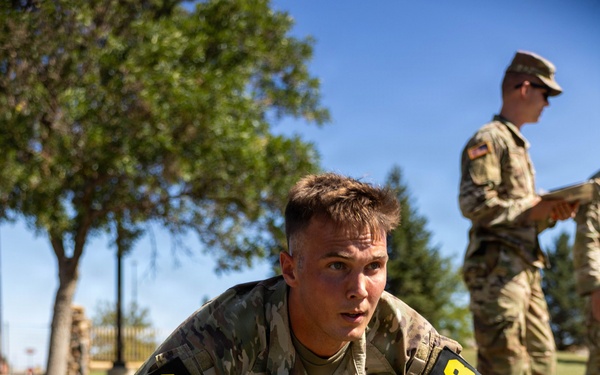 A U.S. Army soldier identified by name tape as 'CARDA' and wearing number '13' bib patches on both shoulders performs push-ups on a grass field. A second uniformed soldier stands in the background holding a clipboard or tablet, apparently observing and scoring the performance. The setting appears to be an outdoor physical fitness test or military competition.