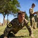 A U.S. Army soldier identified by name tape as 'CARDA' and wearing number '13' bib patches on both shoulders performs push-ups on a grass field. A second uniformed soldier stands in the background holding a clipboard or tablet, apparently observing and scoring the performance. The setting appears to be an outdoor physical fitness test or military competition.