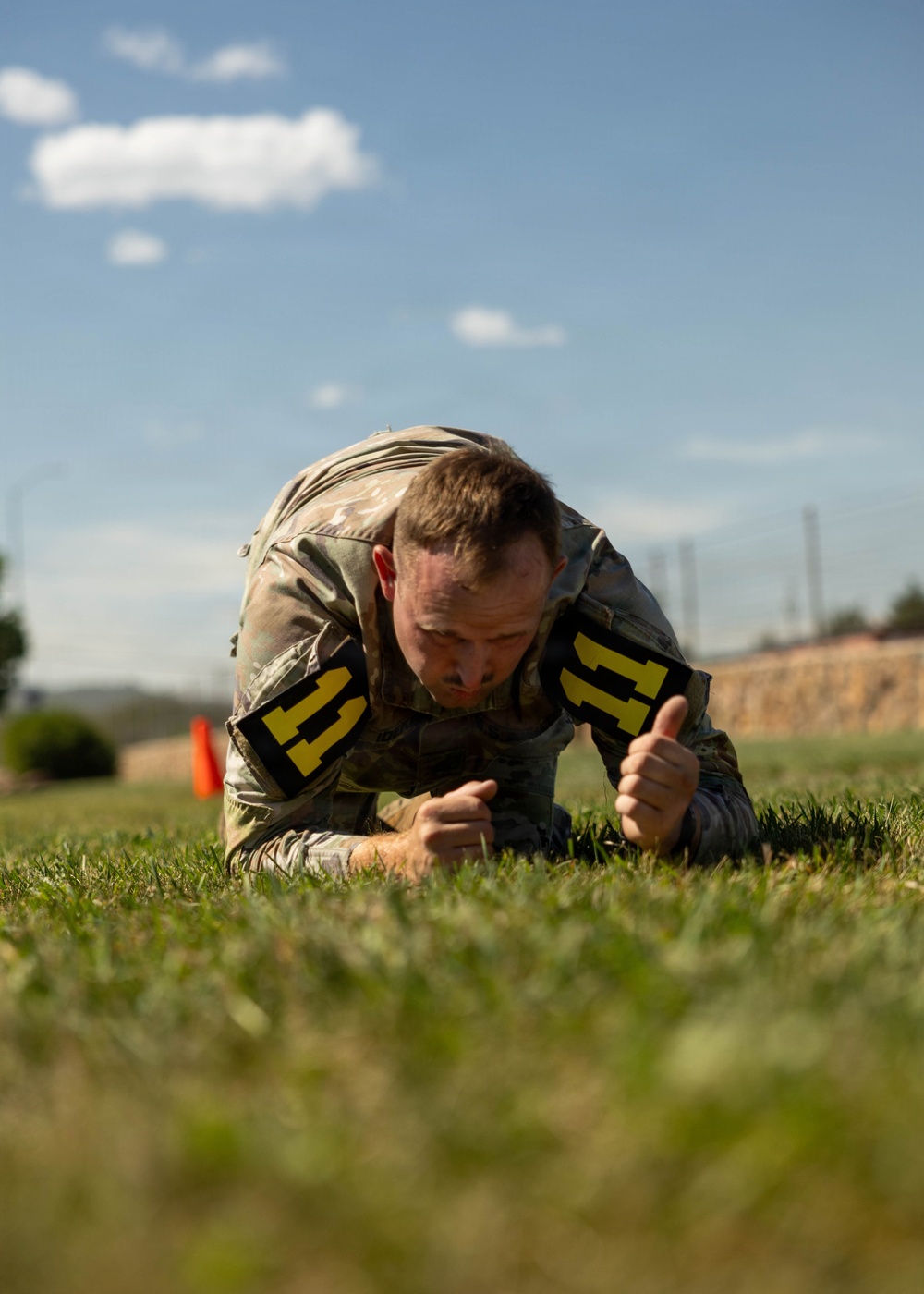 A soldier in camouflage uniform is performing a low crawl exercise on a grass field, wearing number '11' bibs on both arms. The image appears to be taken during a military fitness competition or training event under clear blue skies. The soldier is in a prone position close to the ground, demonstrating focused physical effort.