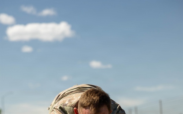 A soldier in camouflage uniform is performing a low crawl exercise on a grass field, wearing number '11' bibs on both arms. The image appears to be taken during a military fitness competition or training event under clear blue skies. The soldier is in a prone position close to the ground, demonstrating focused physical effort.