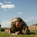 A soldier in camouflage uniform is performing a low crawl exercise on a grass field, wearing number '11' bibs on both arms. The image appears to be taken during a military fitness competition or training event under clear blue skies. The soldier is in a prone position close to the ground, demonstrating focused physical effort.