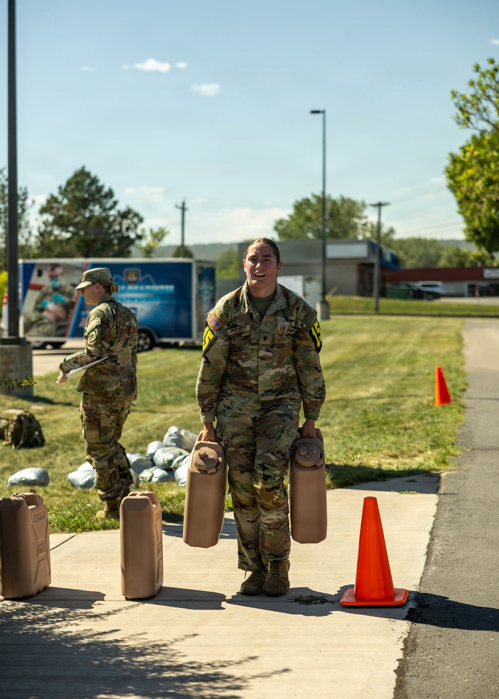 A U.S. Army soldier identified by name tape as 'PUCKETT' carries two military-style water/fuel canisters (jerry cans) along a paved walkway during what appears to be a training exercise or logistics activity. A second soldier in the background holds a clipboard or document. Multiple additional canisters are visible on the ground along the path, marked by orange traffic cones.