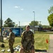 A U.S. Army soldier identified by name tape as 'PUCKETT' carries two military-style water/fuel canisters (jerry cans) along a paved walkway during what appears to be a training exercise or logistics activity. A second soldier in the background holds a clipboard or document. Multiple additional canisters are visible on the ground along the path, marked by orange traffic cones.