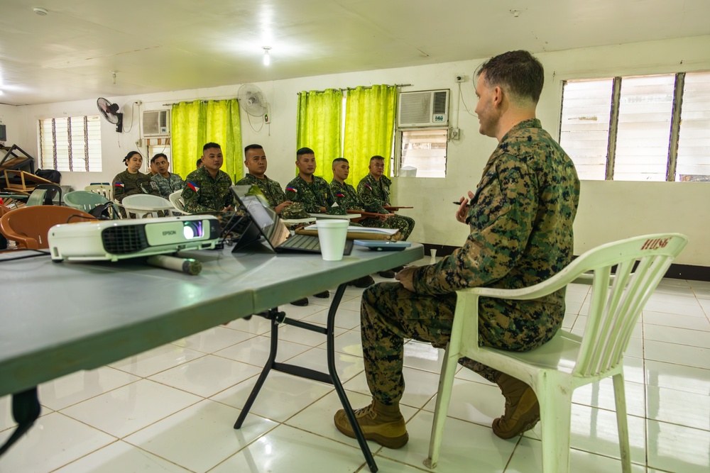A military briefing or meeting is taking place in a room with soldiers seated at tables. One individual in US Marine Corps MARPAT digital camouflage sits facing a group of soldiers wearing Philippine Armed Forces uniforms with visible Philippine flag patches. An Epson projector and laptop are set up on the table, suggesting a formal presentation or training session.