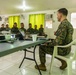 A military briefing or meeting is taking place in a room with soldiers seated at tables. One individual in US Marine Corps MARPAT digital camouflage sits facing a group of soldiers wearing Philippine Armed Forces uniforms with visible Philippine flag patches. An Epson projector and laptop are set up on the table, suggesting a formal presentation or training session.