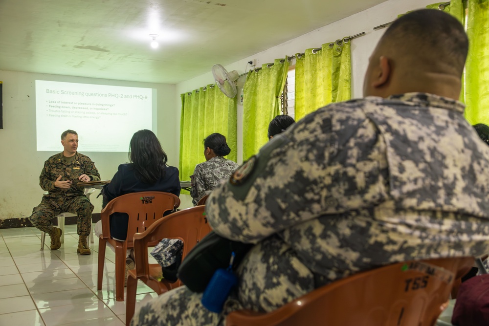 A military briefing or training session is taking place in a room where a U.S. Marine in woodland camouflage uniform is presenting to a small group of attendees, some of whom are wearing military uniforms. A projected slide on the wall displays 'Basic Screening Questions PHQ-2 and PHQ-9,' indicating this is a mental health screening or awareness training. The audience is seated in orange plastic chairs facing the presenter.
