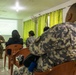 A military briefing or training session is taking place in a room where a U.S. Marine in woodland camouflage uniform is presenting to a small group of attendees, some of whom are wearing military uniforms. A projected slide on the wall displays 'Basic Screening Questions PHQ-2 and PHQ-9,' indicating this is a mental health screening or awareness training. The audience is seated in orange plastic chairs facing the presenter.