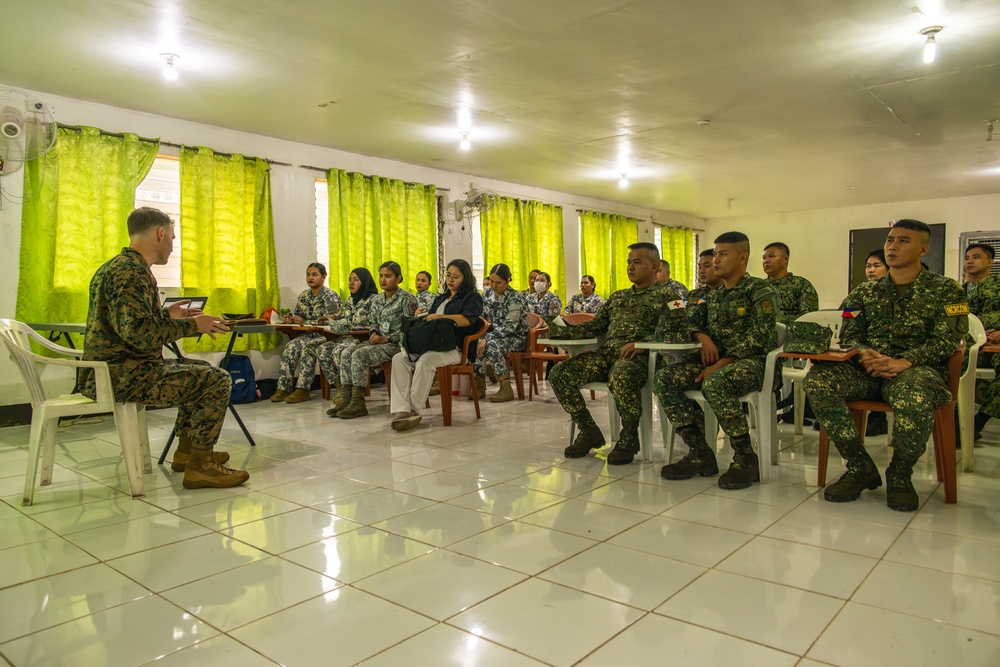 A military briefing or training session is taking place in a room with bright green curtains. One individual in US Marine Corps camouflage uniform sits facing a group of attendees that includes personnel in two different military uniforms and at least one civilian. The setting appears to be a classroom or meeting room in a tropical or Southeast Asian location.