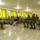 A military briefing or training session is taking place in a room with bright green curtains. One individual in US Marine Corps camouflage uniform sits facing a group of attendees that includes personnel in two different military uniforms and at least one civilian. The setting appears to be a classroom or meeting room in a tropical or Southeast Asian location.