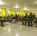 A military briefing or training session is taking place in a room with bright green curtains. One individual in US Marine Corps camouflage uniform sits facing a group of attendees that includes personnel in two different military uniforms and at least one civilian. The setting appears to be a classroom or meeting room in a tropical or Southeast Asian location.