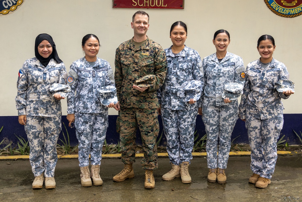 Six military personnel pose for a group photo in front of a wall bearing the word 'SCHOOL' and organizational insignia. Five individuals wear blue digital camouflage uniforms with 'COAST GUARD' patches, consistent with the Philippine Coast Guard, while one individual in the center wears U.S. Marine Corps MARPAT woodland camouflage. All six are holding camouflage covers (patrol caps), suggesting a ceremonial exchange or presentation.
