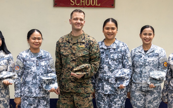 Six military personnel pose for a group photo in front of a wall bearing the word 'SCHOOL' and organizational insignia. Five individuals wear blue digital camouflage uniforms with 'COAST GUARD' patches, consistent with the Philippine Coast Guard, while one individual in the center wears U.S. Marine Corps MARPAT woodland camouflage. All six are holding camouflage covers (patrol caps), suggesting a ceremonial exchange or presentation.