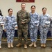 Six military personnel pose for a group photo in front of a wall bearing the word 'SCHOOL' and organizational insignia. Five individuals wear blue digital camouflage uniforms with 'COAST GUARD' patches, consistent with the Philippine Coast Guard, while one individual in the center wears U.S. Marine Corps MARPAT woodland camouflage. All six are holding camouflage covers (patrol caps), suggesting a ceremonial exchange or presentation.