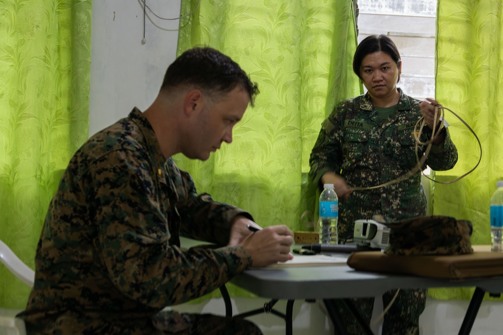 Two military personnel in camouflage uniforms are engaged in what appears to be a training or administrative session indoors. One individual, seated at a table, is writing on a document, while the other, standing, holds what appears to be tubing or cordage. The setting includes a table with water bottles and equipment, against a backdrop of green curtains.