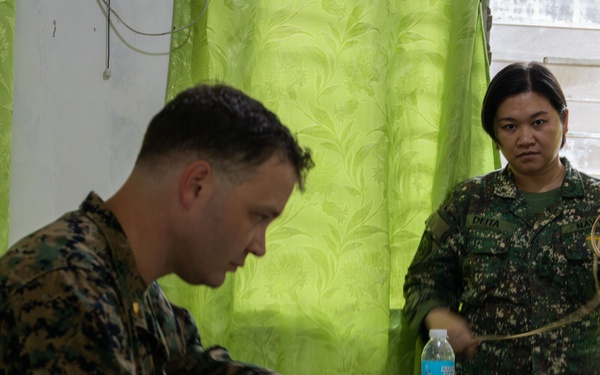 Two military personnel in camouflage uniforms are engaged in what appears to be a training or administrative session indoors. One individual, seated at a table, is writing on a document, while the other, standing, holds what appears to be tubing or cordage. The setting includes a table with water bottles and equipment, against a backdrop of green curtains.