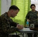 Two military personnel in camouflage uniforms are engaged in what appears to be a training or administrative session indoors. One individual, seated at a table, is writing on a document, while the other, standing, holds what appears to be tubing or cordage. The setting includes a table with water bottles and equipment, against a backdrop of green curtains.