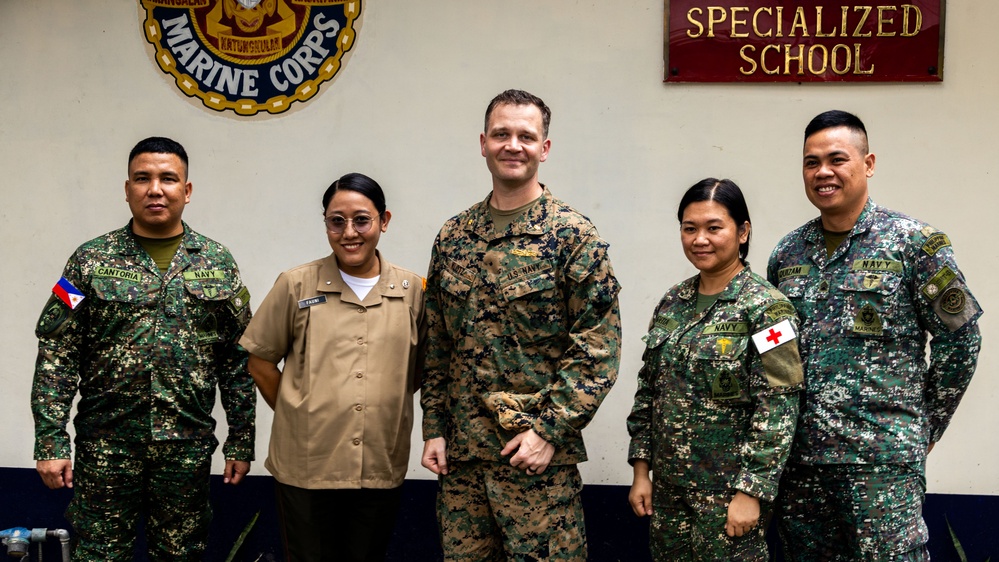 Five military personnel pose for a group photo in front of a 'Specialized School' sign and a Philippine Marine Corps emblem. The group includes members from both the Philippine Marine Corps/Navy and the U.S. Navy/Marine Corps, suggesting a joint military engagement or training event. The setting appears to be inside a Philippine military facility.