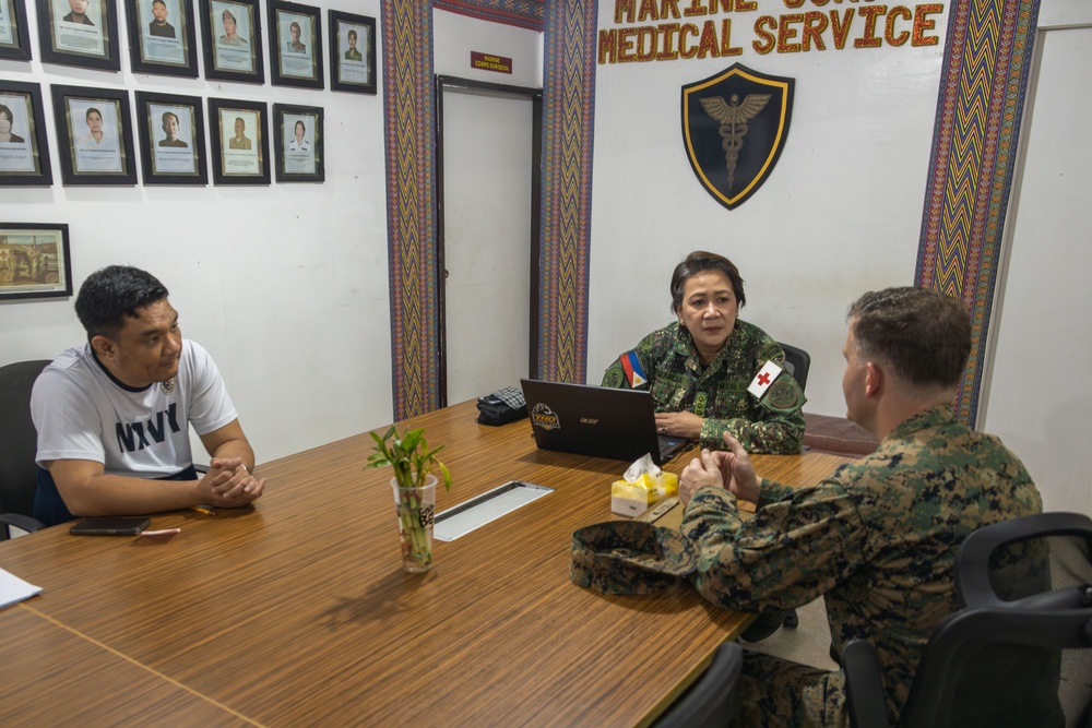 Three individuals are seated around a conference table in a Marine Corps Medical Service office. The meeting involves two military personnel in camouflage uniforms and one civilian in a white t-shirt. The setting appears to be an official military medical facility, likely in the Philippines based on visible insignia.