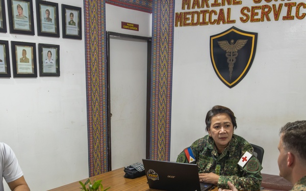 Three individuals are seated around a conference table in a Marine Corps Medical Service office. The meeting involves two military personnel in camouflage uniforms and one civilian in a white t-shirt. The setting appears to be an official military medical facility, likely in the Philippines based on visible insignia.