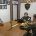 Three individuals are seated around a conference table in a Marine Corps Medical Service office. The meeting involves two military personnel in camouflage uniforms and one civilian in a white t-shirt. The setting appears to be an official military medical facility, likely in the Philippines based on visible insignia.