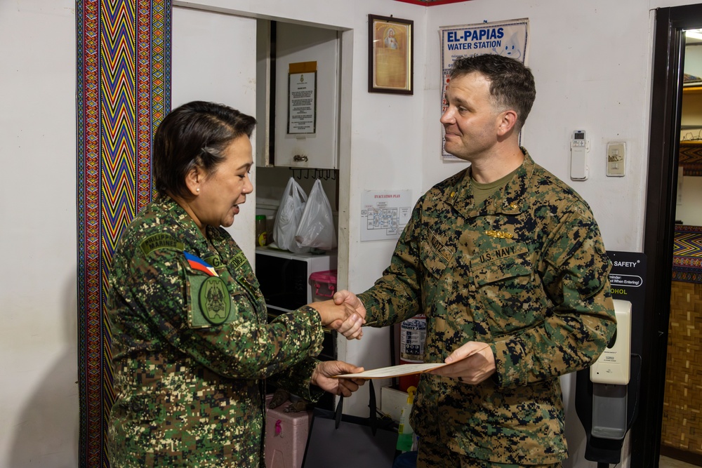 Two military personnel are shaking hands in what appears to be an indoor facility. The individual on the left wears a Philippine Armed Forces uniform with a Philippine flag patch, while the individual on the right wears a U.S. Navy digital camouflage uniform. The U.S. Navy servicemember is holding what appears to be a document or certificate, suggesting a formal exchange or presentation.