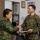 Two military personnel are shaking hands in what appears to be an indoor facility. The individual on the left wears a Philippine Armed Forces uniform with a Philippine flag patch, while the individual on the right wears a U.S. Navy digital camouflage uniform. The U.S. Navy servicemember is holding what appears to be a document or certificate, suggesting a formal exchange or presentation.