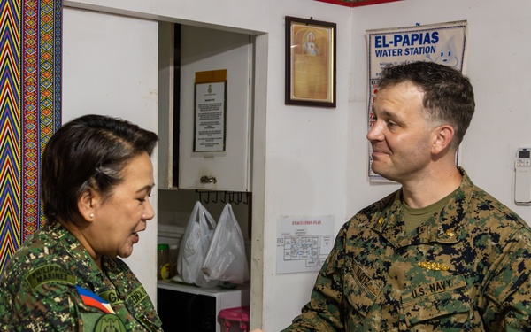 Two military personnel are shaking hands in what appears to be an indoor facility. The individual on the left wears a Philippine Armed Forces uniform with a Philippine flag patch, while the individual on the right wears a U.S. Navy digital camouflage uniform. The U.S. Navy servicemember is holding what appears to be a document or certificate, suggesting a formal exchange or presentation.