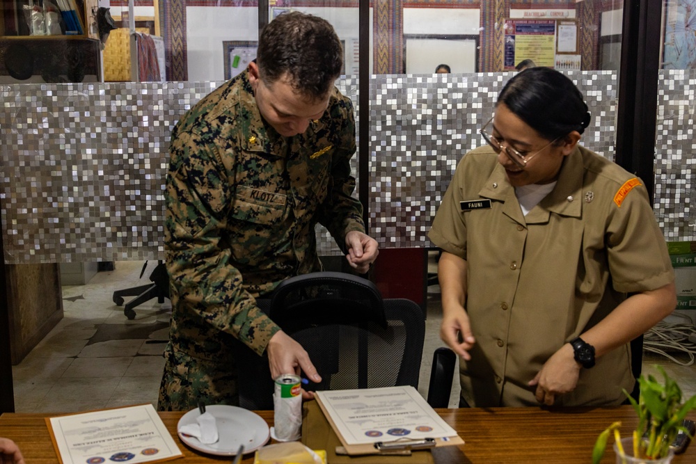Two military personnel, one in US Marine Corps digital camouflage uniform and one in a tan/khaki uniform, are standing at a desk reviewing documents. The Marine, identified by name tape as 'KLOTZ,' appears to be presenting or signing certificates alongside the other service member identified as 'FAUNI.' The setting appears to be a military office or administrative area.